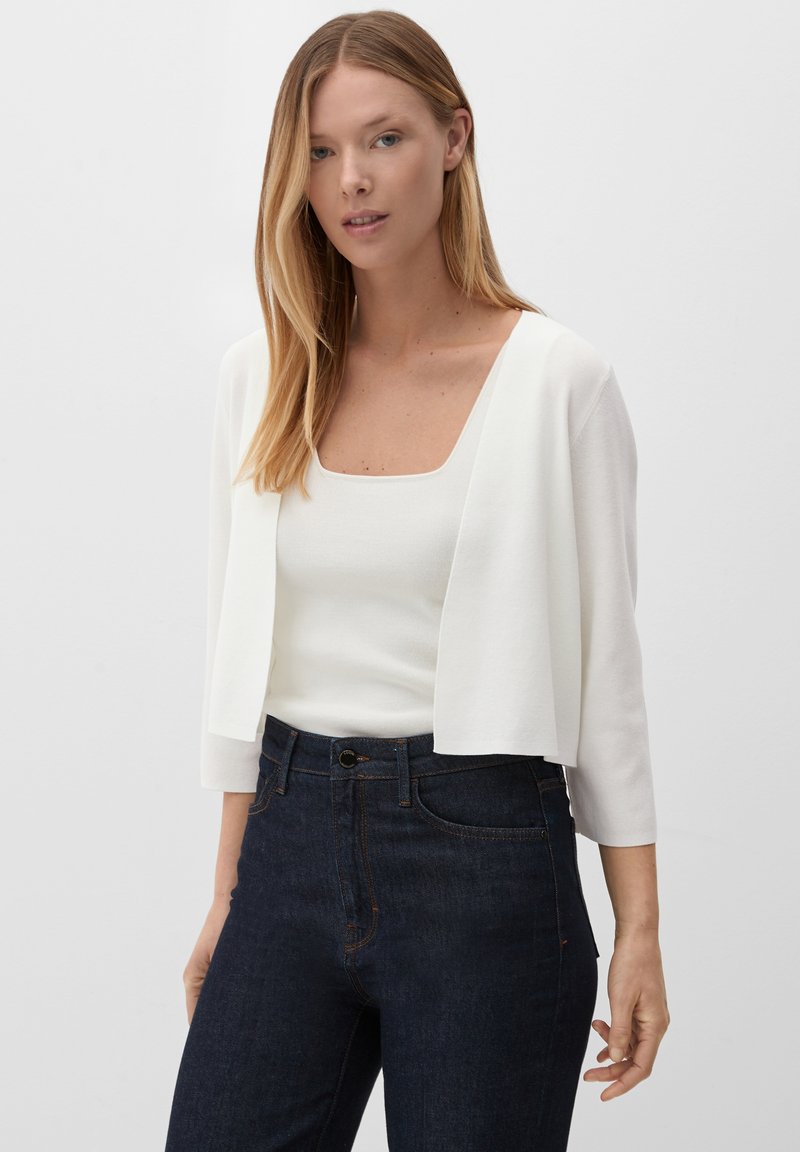 Witte cropped cardigan over een aansluitend wit topje, gecombineerd met donkere spijkerbroeken. De cardigan heeft driekwart mouwen en een vierkante halslijn.