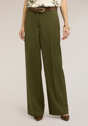 Kvinde iført olivengrønne bukser med vide ben, brun læderbælte, blomsterbluse, gyldne armbånd og brune spidse sko.