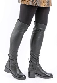Botas negras de cuero por encima de la rodilla con una textura suave, punta redondeada, tacón cuadrado y detalle de cremallera lateral. Combinadas con una falda estampada.