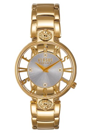 Reloj de pulsera Versus Versace en tono dorado con esfera plateada, letras caladas que forman "VERSUS", cuatro marcadores de diamante y detalles de cabeza de león en la pulsera.