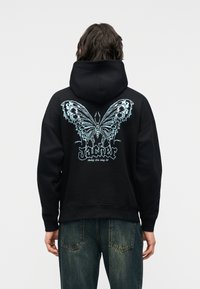 Oseba, ki nosi črno kapuco z veliko podrobno modro metuljasto grafiko in stiliziranim besedilom "Jacker" na hrbtu, oblečena v modre jeans hlače.