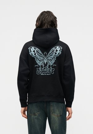 Personne portant un sweat à capuche noir avec un grand papillon bleu détaillé et un texte stylisé « Jacker » dans le dos, associé à un jean bleu.