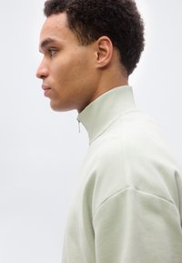 Νέος άνδρας με ανοιχτό γκρι πουλόβερ με γιακά μισής ζώνης, στραμμένος προς τα αριστερά με φόντο ένα απλό λευκό υπόβαθρο.