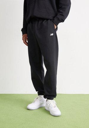 Osoba nosząca czarne spodnie dresowe New Balance i białe buty Nike stoi na zielonej podłodze na tle gładkiej jasnej ściany.
