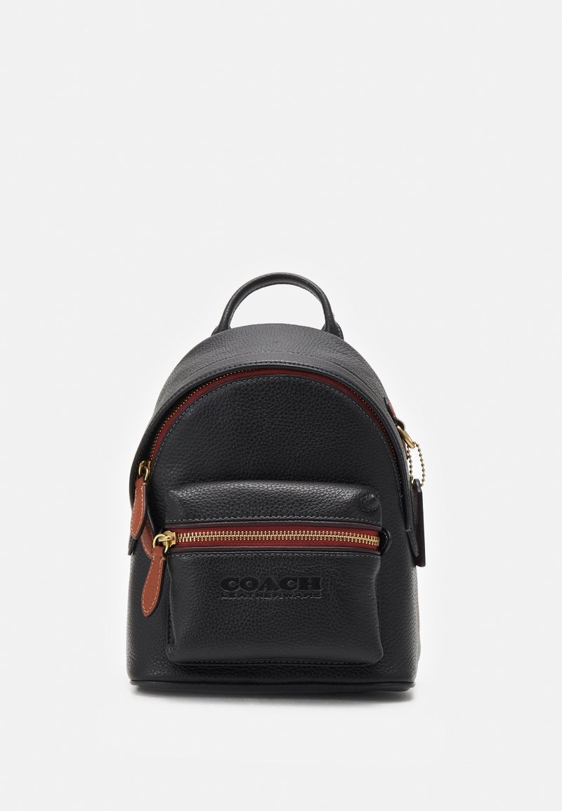 Petit sac à dos en cuir noir avec poche zippée à l'avant et tirettes de fermeture éclair marron sur fond uni clair.
