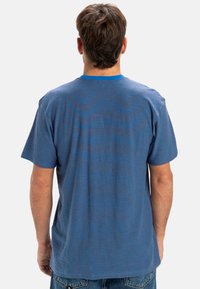 Modré pruhované tričko s krátkým rukávem, pohled zezadu. Materiál vypadá měkce, s horizontálními modrými a hnědými pruhy na látce.