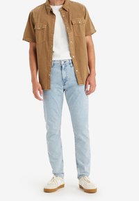 Man som bär ljusblå jeans, vit t-shirt, öppen brun kortärmad skjorta med knappar och beige sneakers med vita snören.