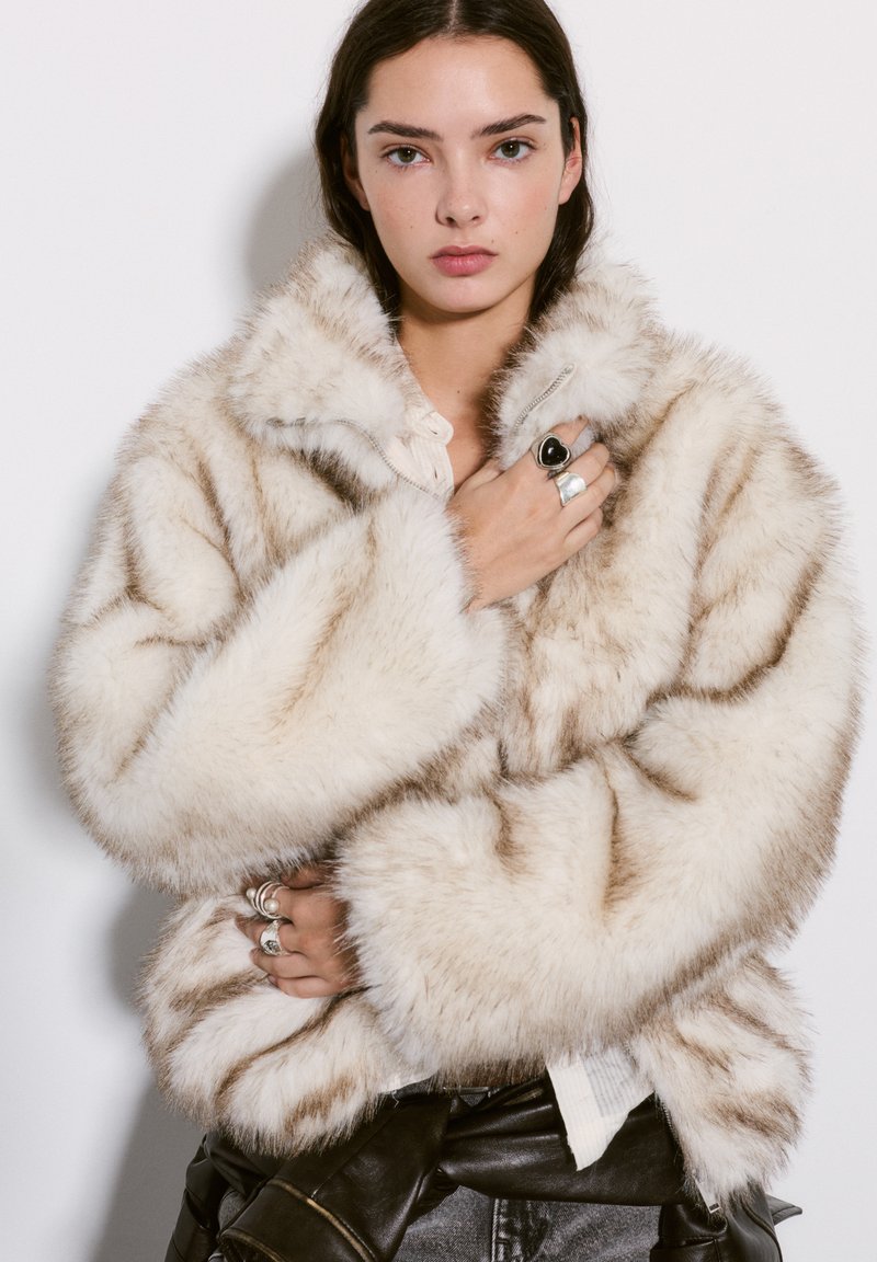 Cream faux fur jacket with a fluffy texture, wide collar, long sleeves, and subtle brown stripes. Zipper closure and oversized fit.