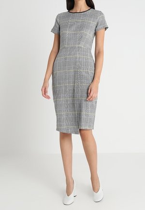 Kobieta ubrana w szarą sukienkę w kratę, o długości do kolan i krótkim rękawie, w połączeniu z białymi zamkniętymi butami, stojąca na jednolitym tle.