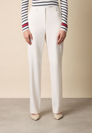 Femme portant un pantalon blanc droit, des chaussures beiges à bout pointu, et un haut à manches longues rayé avec des accents rouges sur les manches.