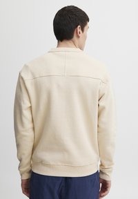 Sudadera de color beige con cuello redondo acanalado, mangas con puños y dobladillo recto. Presenta un mínimo detalle de costura en la parte posterior.