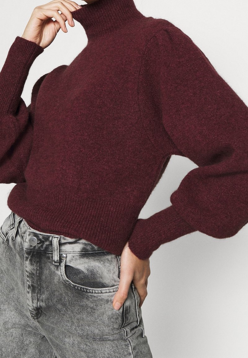 Pull en maille bordeaux avec un col montant et des manches bouffantes, associé à un pantalon en denim gris. Le pull présente une texture côtelée aux poignets et à l'ourlet.