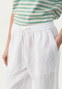 Pantalons en lin blanc avec taille élastique et cordon de serrage, dotés de deux poches latérales et d'une texture lisse, associés à un haut rayé vert et crème.