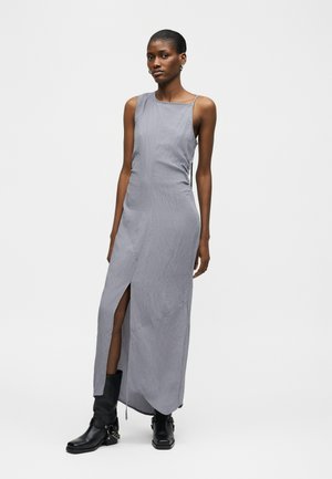 Γυναίκα που φοράει αμάνικο γκρι μακρύ φόρεμα με σκίσιμο μπροστά και μαύρα μποτάκια μέχρι τη μέση της γάμπας, στέκεται μπροστά σε ένα απλό φωτεινό φόντο.