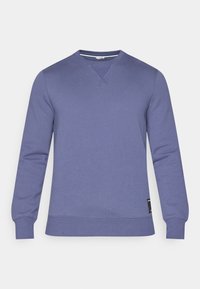 Sweatshirt à col rond violet en tissu doux. Il présente des manches longues, des poignets côtelés et un détail cousu au niveau du col. Accent avec une étiquette noire.