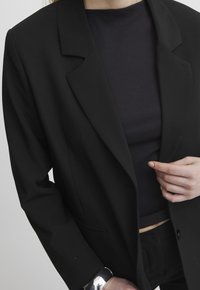 Osoba z czarnym żakietem noszącym szary, prążkowany top, z jedną ręką poprawiającą żakiet oraz odblaskową srebrną bransoletą na nadgarstku.