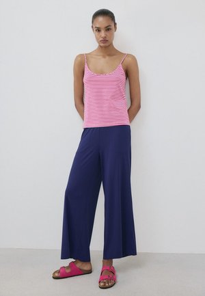 Femme portant un débardeur rayé rose et blanc, un pantalon large bleu marine et des sandales roses à boucle, debout contre un mur blanc.