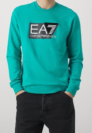 Sweat-shirt à manches longues bleu sarcelle avec logo EA7 Emporio Armani en noir et blanc, porté par une personne les mains dans les poches d'un pantalon noir.