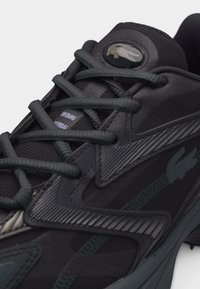 Κοντινή εικόνα μαύρου αθλητικού παπουτσιού με υφασμένα κορδόνια, λεπτομερείς ραφές και στρογγυλό λογότυπο με τον κροκόδειλο στη γλώσσα.