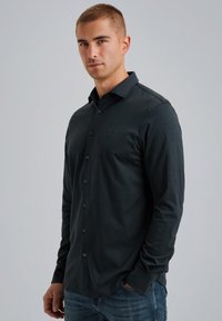 Tummanvihreä pitkähihainen nappikauluspaita, joka on valmistettu sileästä kankaasta. Paidassa on avonainen kaulus, etupaneeli ja hillitty logoyksityiskohta rinnassa.