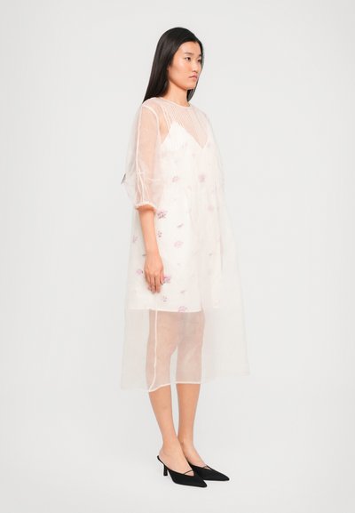 Áttetsző pasztell rózsaszín ruha hímzett virágmintákkal, laza fazonban, térd hosszúságú és puffos ujjal. Fekete magassarkú mamuszokkal egészítve.