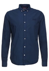 Námořnická modrá košile s dlouhým rukávem a knoflíky, s špičatým límcem, bílými knoflíky a malým vyšitým logem na hrudi. Bavlněný materiál.