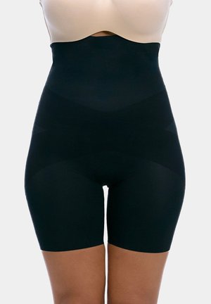 Woman wearing beige bra and high-waisted black shaping shorts that extend to mid-thigh on a plain light background.