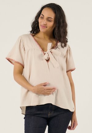 Femme enceinte en blouse beige avec détail nœud, tenant doucement son ventre, regardant vers le bas avec un doux sourire, portant un jean foncé.