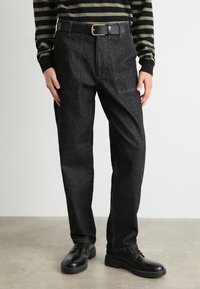 Pantaloni din denim negru cu o croială relaxată, dotati cu buzunare frontale, un curea neagră cu o cataramă aurie și asortați cu pantofi negri.