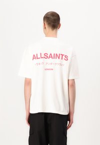 T-shirt blanc à manches courtes avec le texte "ALLSAINTS" en rose bien visible et des caractères japonais au dos. Présente une coupe décontractée et un col rond.
