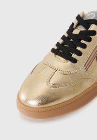 Sneaker in pelle dorata con lacci neri, finitura testurizzata e suola in gomma. Presenta una cerniera laterale e dettagli di cucitura a contrasto.