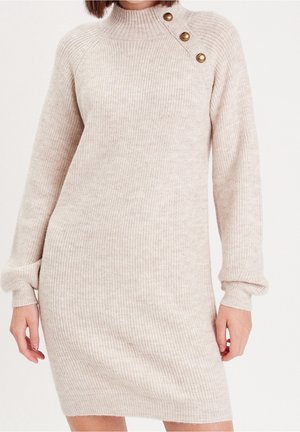 Robe beige en maille côtelée à manches longues avec col montant et trois boutons dorés sur l'épaule gauche, portée par une personne debout.