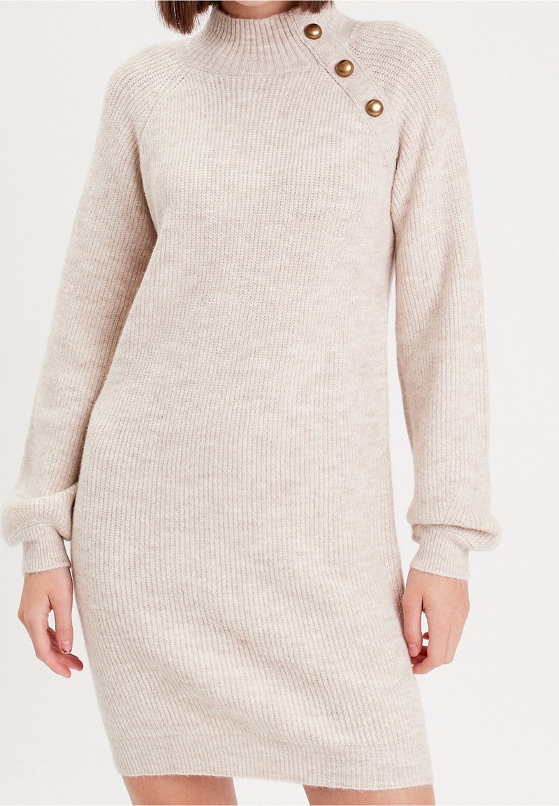 Robe beige en maille côtelée à manches longues avec col montant et trois boutons dorés sur l'épaule gauche, portée par une personne debout.
