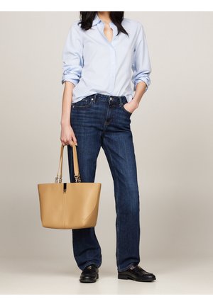 Chemise boutonnée bleu clair, jean droit bleu foncé, et un sac cabas en cuir beige avec une anse en chaîne argentée. Des chaussures noires complètent la tenue.