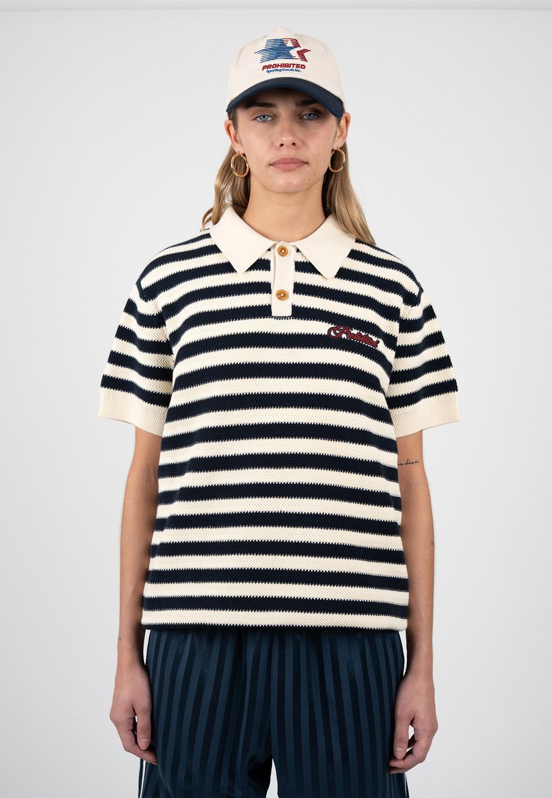 Femme portant un polo à manches courtes rayé bleu marine et blanc, un pantalon rayé bleu marine, une casquette beige et bleu marine, et des boucles d'oreilles créoles en or.
