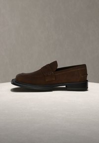 Mocassin en cuir marron avec semelle noire, coutures détaillées et bride à boucle, posé sur une surface claire devant un fond gris.