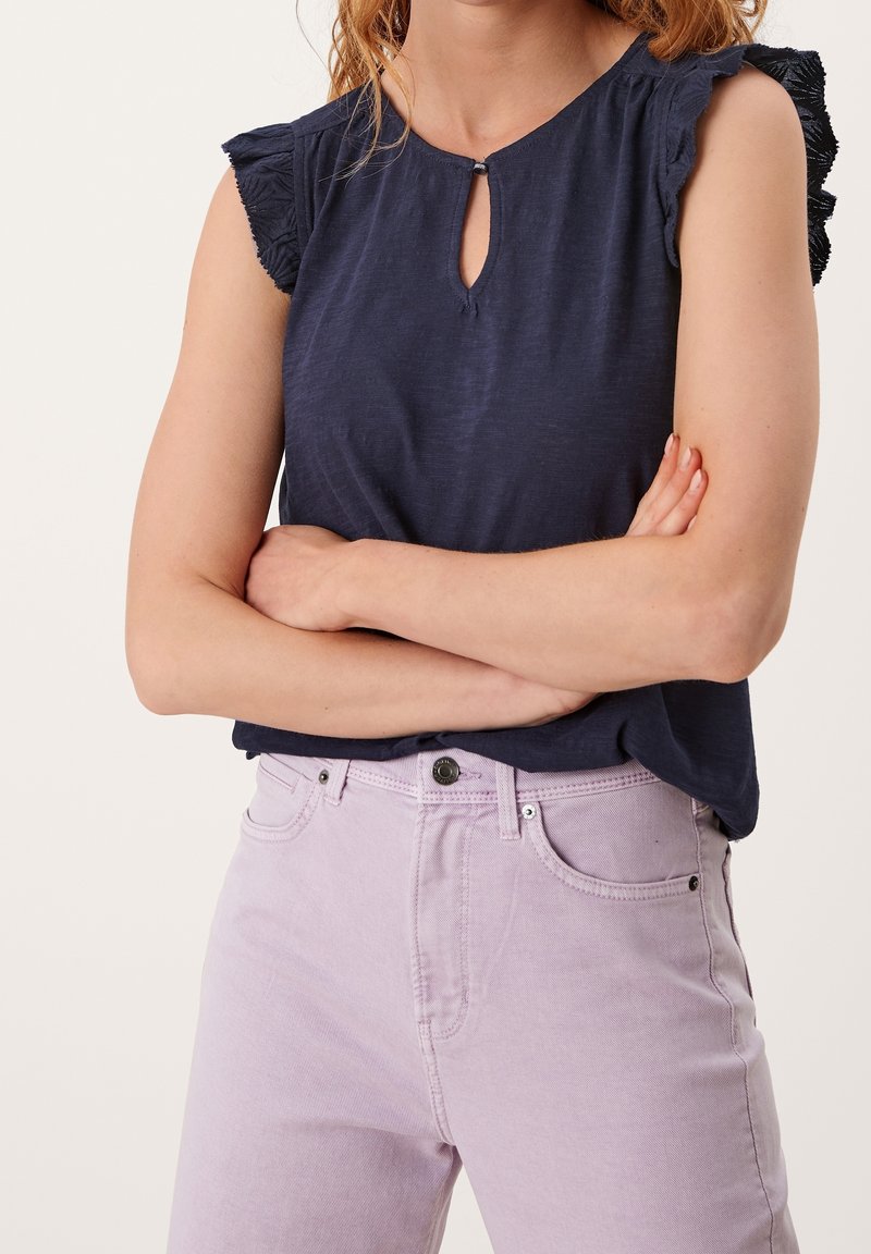 Top senza maniche color blu navy con scollatura a chiave, caratterizzato da spalle arricciate. Abbinato a jeans leggeri color lavanda, con una texture liscia.
