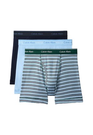 Męski model stoi na boso, nosząc czarne bokserki Calvin Klein na jednolitym szarym tle z jedną ręką za plecami.
