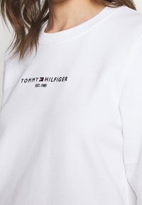 Sudadera blanca de algodón con cuello acanalado. Presenta el logotipo bordado en negro "TOMMY HILFIGER" y "EST. 1985" en colores contrastantes.