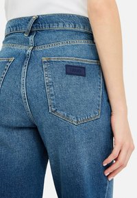 Jeans de tiro alto de mezclilla azul claro con un bolsillo trasero que presenta una etiqueta de cuero azul oscuro. La tela parece texturizada con sutiles detalles de costura.