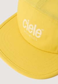 Sárga sapka, textúrált felülettel és lapos szélességgel, a "ciele ATHLETICS" felirattal fehér színben az elején.