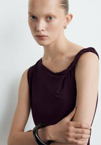 Vestido sin mangas color granate con detalle torcido en el hombro, combinado con una pulsera negra. La modelo tiene piel suave y ojos azules, con el cabello peinado hacia atrás.