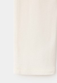 Gros plan d’un tissu couleur crème avec un ourlet cousu visible sur le bord inférieur, sur un fond clair uni.