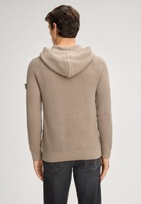 Hoodie en tricot beige avec un motif texturé, des poignets et un ourlet côtelés, et une couture centrale le long du dos. Porté avec un jean en denim noir.