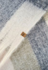 Uldhalstørklæde med et geometrisk mønster i lys grå, beige, lys blå og hvid, med et lille mærket lædertag syet ind i kanten.