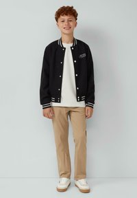Zwarte varsityjack met witte accenten, witte t-shirt, beige broek en witte sneakers met rubberen zolen. Korte gekrulde haarstijl.