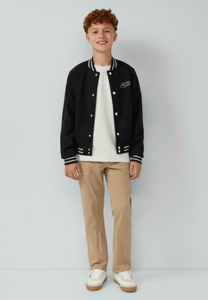 Zwarte varsityjack met witte accenten, witte t-shirt, beige broek en witte sneakers met rubberen zolen. Korte gekrulde haarstijl.