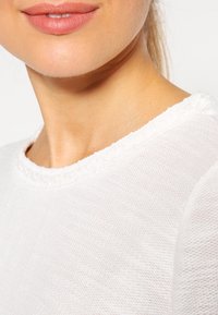Bílý texturovaný pletený top s kulatým výstřihem a dekorativním lemováním kolem límce, úzce střižený, aby ukázal detail tkaniny a tvar výstřihu.