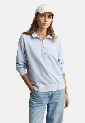 Vrouw die een lichtblauwe trui met kwartrits, lichtblauwe spijkerbroek en een witte gestreepte baseballpet draagt, met één hand in haar zak poseert.