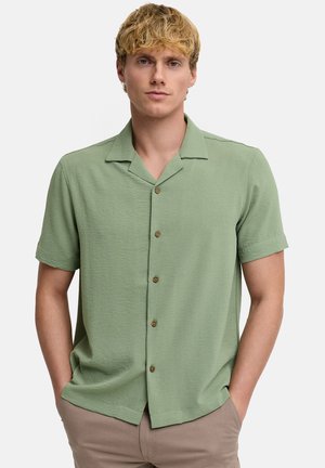 Jeune homme aux cheveux blonds portant une chemise à manches courtes vert clair boutonnée et un pantalon beige, mains dans les poches, faisant face.